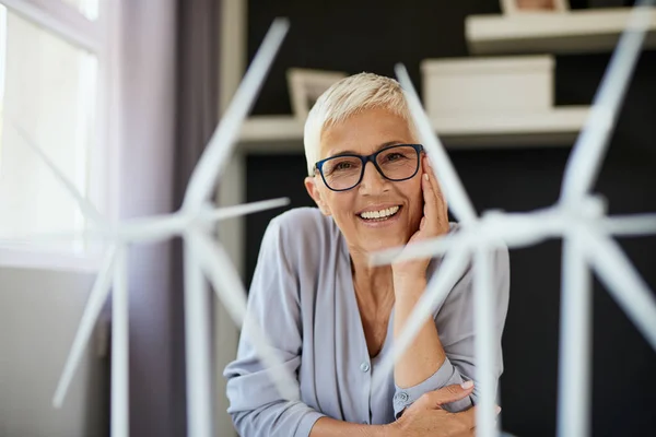 Fröhliche kaukasische Seniorin, die sich auf den Schreibtisch lehnt und in die Kamera blickt. Auf dem Schreibtisch stehen Windmühlenmodelle. Existenzgründungskonzept. — Stockfoto