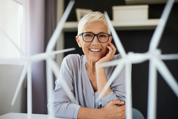 Happy Caucasian senior woman leaning on desk and looking at camera. On desk are windmill models. Start up business concept.