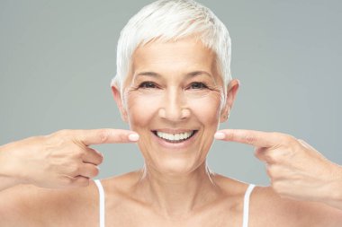 Beautiful Caucasian  smiling senior woman with short grey hair pointing at her teeth and looking at camera. Beauty photography. clipart