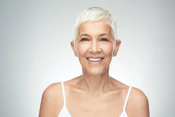 Bella donna anziana sorridente con i capelli corti grigi in posa di fronte a sfondo grigio. Foto di bellezza . — Foto Stock