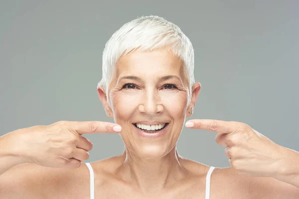 Hermosa mujer mayor caucásica sonriente con el pelo gris corto apuntando a sus dientes y mirando a la cámara. Belleza fotografía . — Foto de Stock