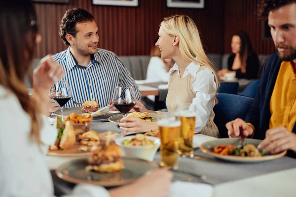 Multietnické přátele, kteří sedí v restauraci, konzumují alkohol, chatují a mají hamburgery na večeři. — Stock fotografie
