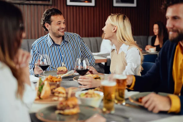 Amigos multiétnicos sentados en el restaurante, bebiendo alcohol, charlando y comiendo hamburguesas para cenar . — Foto de Stock