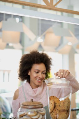 Pembe balıkçı yaka kazak ve kurabiye ile önlük açılış kavanozu ile çekici karışık ırk çalışanı. Pastane içi.