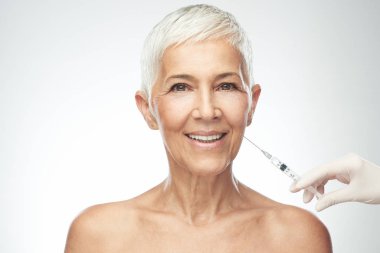 Beautiful smiling Caucasian senior woman standing in front of gray background and taking a shot of anti age product. Beauty photography. clipart