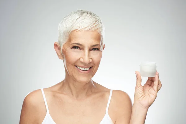 Eine zauberhaft lächelnde kaukasische Seniorin probiert eine neue Anti-Age-Creme aus und blickt in die Kamera. Schönheitsfotografie. — Stockfoto