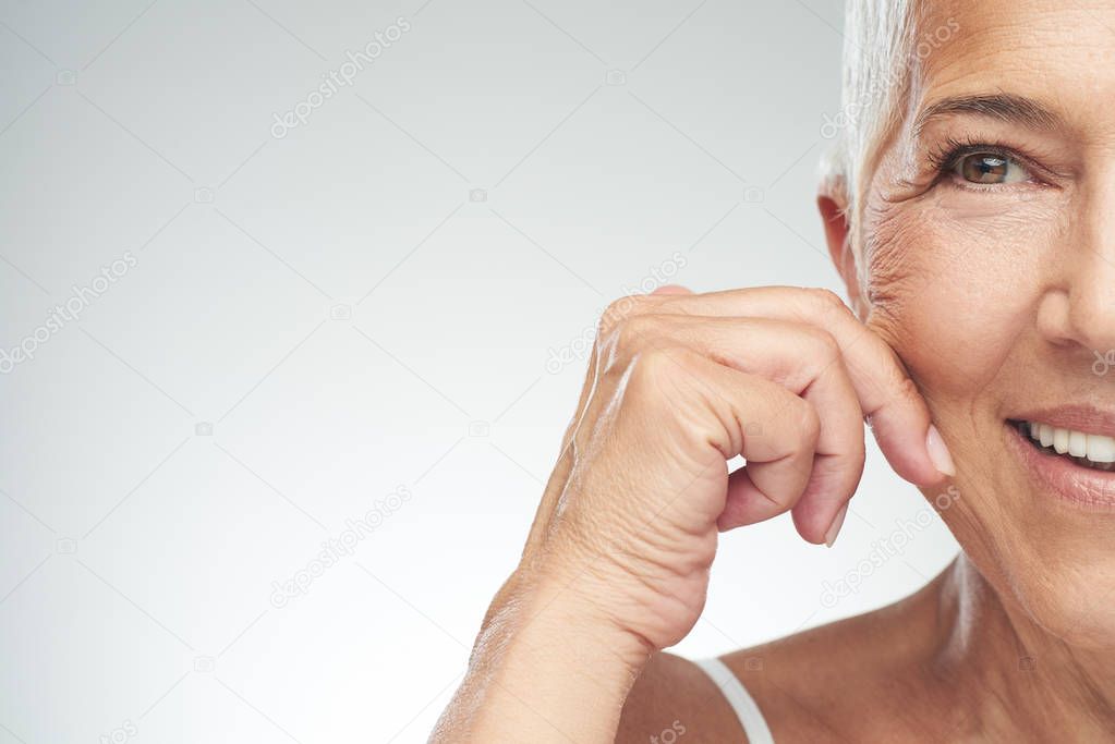 Gorgeous smiling Caucasian senior woman with short gray hair pinching her cheek. Beauty photography.