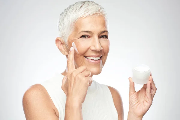 Eine zauberhaft lächelnde kaukasische Seniorin probiert eine neue Anti-Age-Creme aus und blickt in die Kamera. Schönheitsfotografie. — Stockfoto