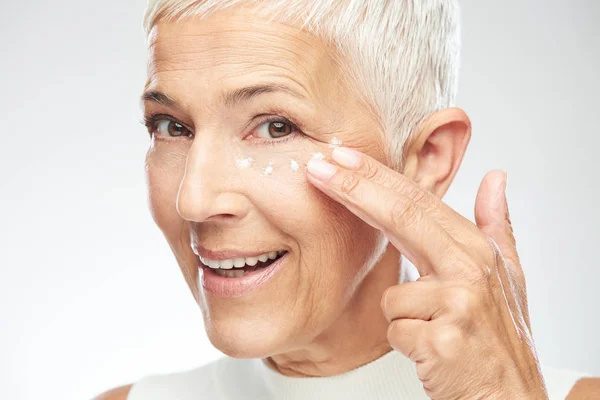 Eine zauberhaft lächelnde kaukasische Seniorin probiert eine neue Anti-Age-Creme aus und blickt in die Kamera. Schönheitsfotografie. — Stockfoto