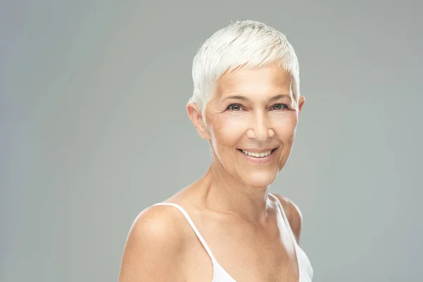 Mooie lachende Senior vrouw met korte grijze haren poseren in de voorkant van de grijze achtergrond. Beauty fotografie. — Stockfoto