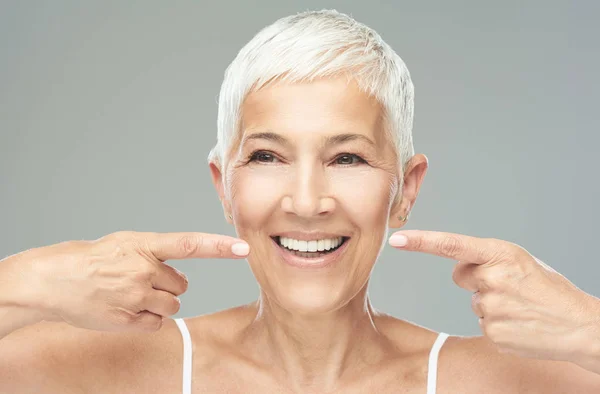 Hermosa mujer mayor caucásica sonriente con el pelo gris corto apuntando a sus dientes y mirando a la cámara. Belleza fotografía . —  Fotos de Stock