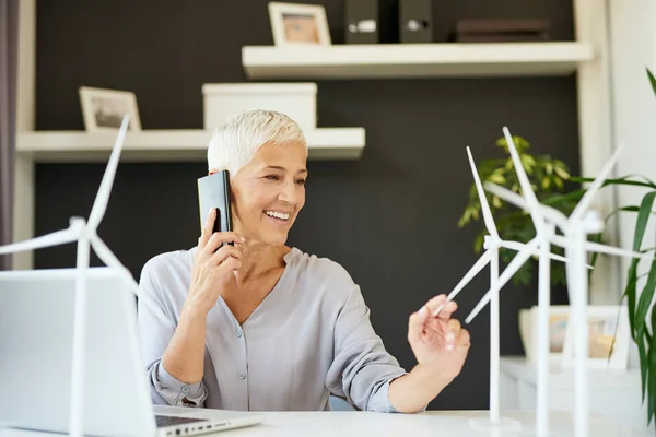 Preciosa mujer mayor caucásica hablando por teléfono y tocando el modelo de molino de viento mientras está sentado en la oficina. Concepto de creación de empresas . — Foto de Stock