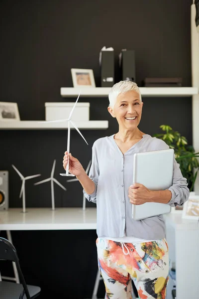 Atractiva sonriente mujer mayor caucásica vestida casual y con el pelo gris corto de pie en la oficina, sosteniendo el ordenador portátil y molino de viento. Concepto sostenible . — Foto de Stock