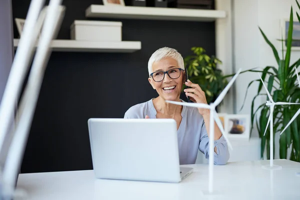 Wanita senior Kaukasia yang cantik berbicara di telepon saat duduk di kantor. Di meja adalah model kincir angin dan laptop.Start up konsep bisnis . — Stok Foto