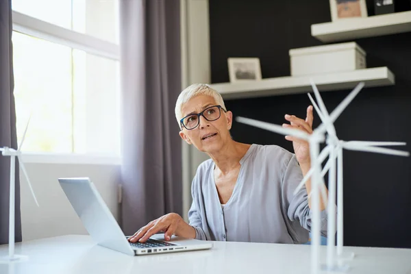 Серьёзная белая старшая женщина пользуется ноутбуком и разговаривает с коллегой во время работы в офисе. На столе находятся ноутбуки и модели ветряной мельницы. Устойчивая концепция . — стоковое фото