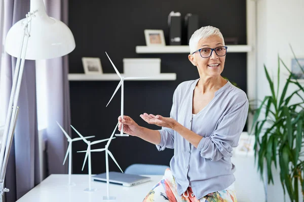 Hermosa mujer mayor caucásica sonriente apoyada en el escritorio, sosteniendo el modelo del molino de viento y hablando con su colega. Concepto sostenible . — Foto de Stock