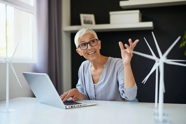 Sonriente mujer mayor caucásica usando el ordenador portátil y hablando con su colega mientras está sentado en la oficina. En el escritorio están los modelos de ordenador portátil y molino de viento. Concepto sostenible . — Foto de Stock