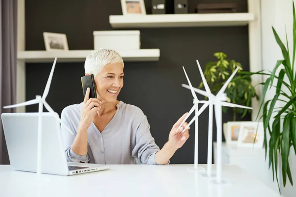 Magnifique femme âgée caucasienne parlant au téléphone et touchant modèle moulin à vent tout en étant assis au bureau. Concept de création d'entreprise . — Photo