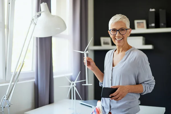 Красивая улыбающаяся белая старшая женщина, стоящая в офисе, держа в руках модель мельницы и смартфон. Концепция стартапа . — стоковое фото