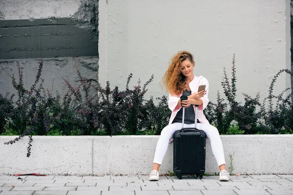 Viajero femenino usando un teléfono inteligente y sosteniendo café mientras está sentado en la pared. Equipaje junto a ella . — Foto de Stock