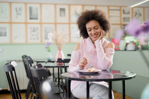 Portret van charmante positieve mixed race vrouw in roze coltrui zitten in zoetwaren en eten gateau. — Stockfoto