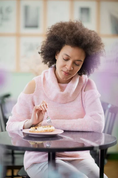 Retrato de encantadora mulher de raça mista positiva em suéter de gola alta rosa sentado em confeitaria e comer gateau . — Fotografia de Stock
