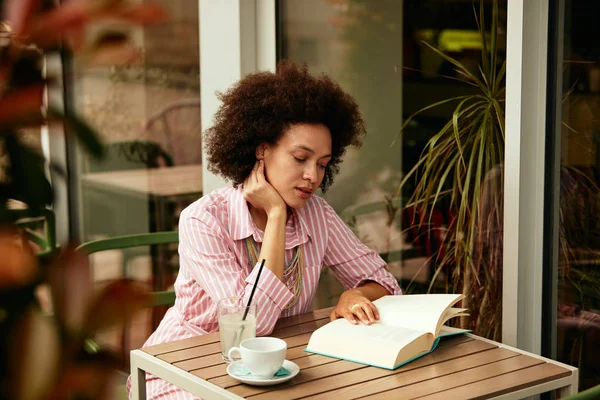 Wspaniała kobieta mieszane rasy w różowe paski sukienka siedzi w kawiarni i czytania książki. — Zdjęcie stockowe