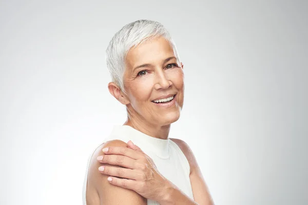 Mooie lachende Senior vrouw met korte grijze haren poseren in de voorkant van de grijze achtergrond. Beauty fotografie. — Stockfoto