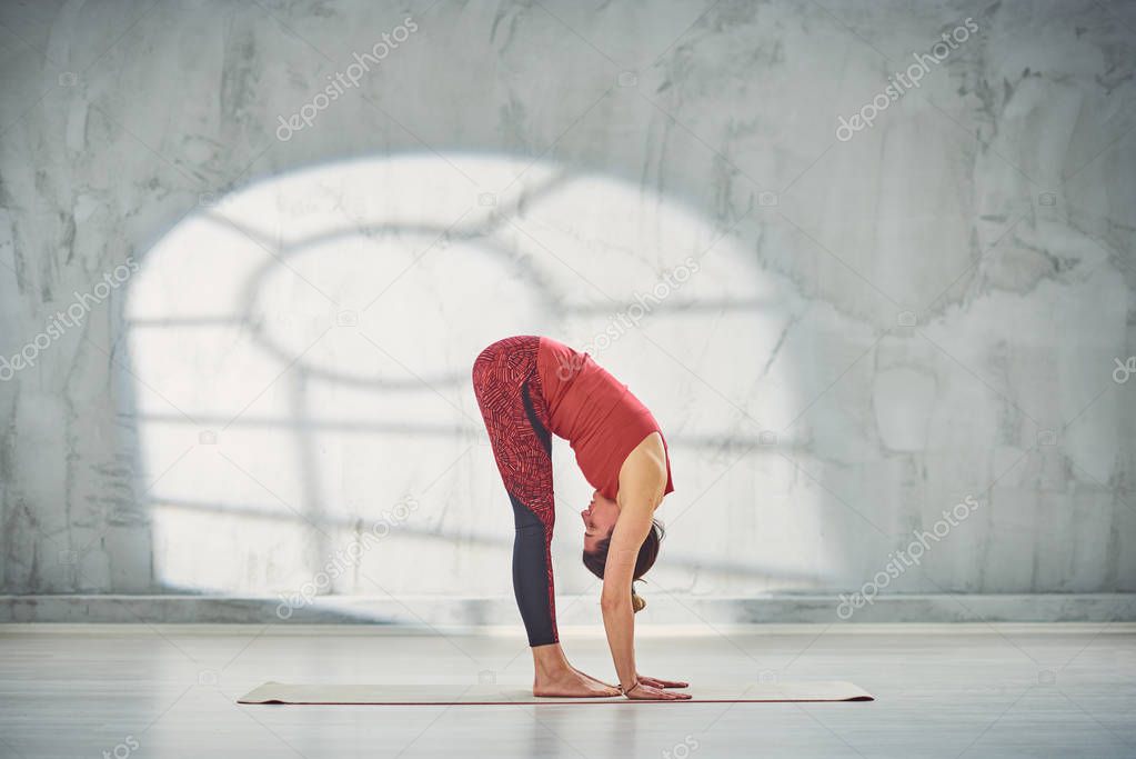 Side view of beautiful Caucasian brunette in red sports wear standing barefoot on the mat in Standing Forward Bend yoga posture.