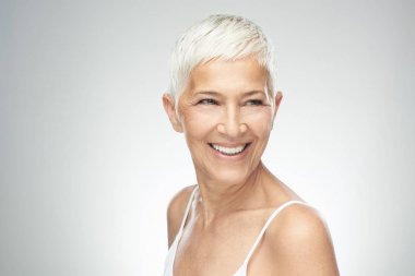 Gri arka plan önünde poz kısa gri saçlı güzel gülümseyen yaşlı kadın. Güzellik fotoğrafçılığı.