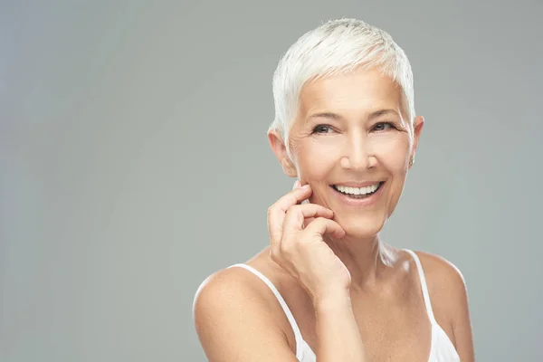 Mooie lachende Senior vrouw met korte grijze haren poseren in de voorkant van de grijze achtergrond. Beauty fotografie. — Stockfoto