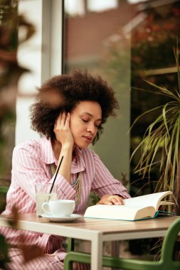Pembe çizgili elbise li muhteşem karışık yarış kadını kafede oturur ken ve kitap okuma.