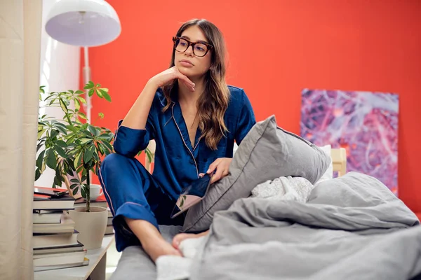 Gorgeous Caucasian brunette with eyeglasses and dressed in blue pajamas sitting on bed in bedroom in the morning and using tablet. — Stock Photo, Image