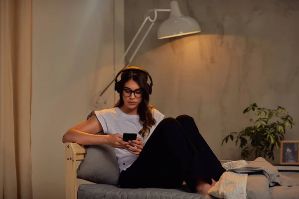 Pretty Caucasian brunette in pajamas and with headphones on head using smart phone late at night while sitting on the bed in bedroom. — Stock Photo, Image