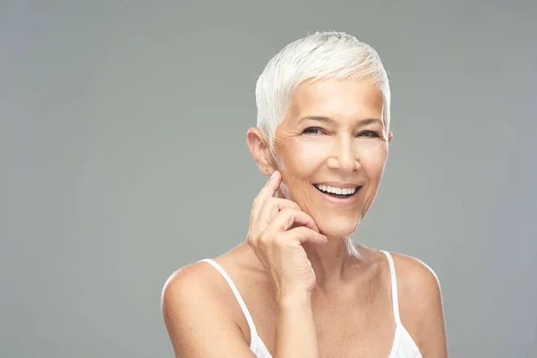 Bella donna anziana sorridente con i capelli corti grigi in posa di fronte a sfondo grigio. Foto di bellezza . — Foto Stock