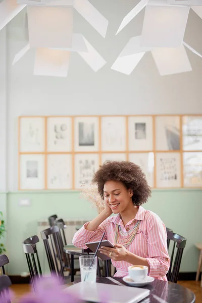 Attraktiv blandad ras kvinna i randig rosa klänning sitter i kafé och använder Tablet. — Stockfoto