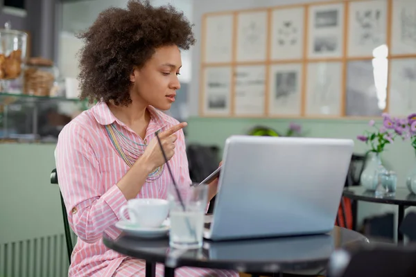 Attraente donna razza mista in abito rosa a righe seduto in caffè e utilizzando tablet . — Foto Stock