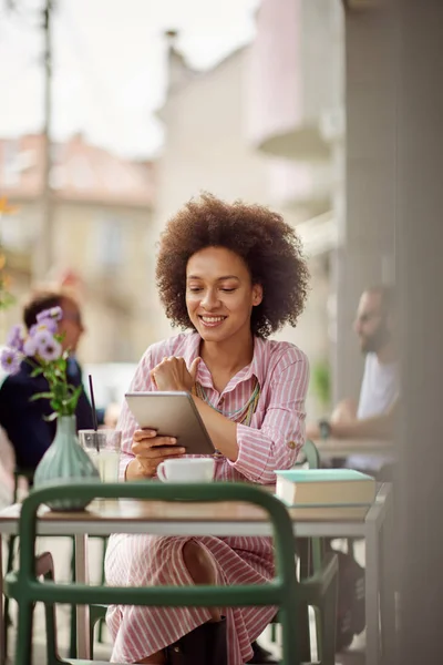 Sorrindo grogeous mulher de raça mista em rosa vestido listrado sentado no café e usando tablet. Na mesa estão xícara de café, livro e limonada . — Fotografia de Stock