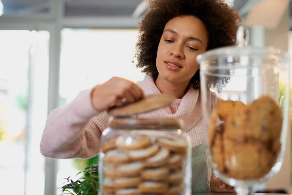 Attractive mixed race employee in pink turtleneck sweater and with apron opening jar with cookies. Pastry shop interior.