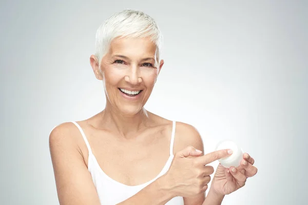 Eine zauberhaft lächelnde kaukasische Seniorin probiert eine neue Anti-Age-Creme aus und blickt in die Kamera. Schönheitsfotografie. — Stockfoto