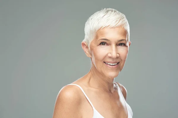 Hermosa mujer mayor sonriente con el pelo gris corto posando delante de fondo gris. Belleza fotografía . —  Fotos de Stock