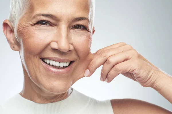 Preciosa mujer mayor caucásica sonriente con el pelo gris corto pellizcando su mejilla. Belleza fotografía . —  Fotos de Stock