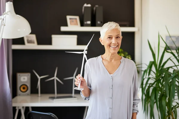 Hermosa mujer mayor caucásica sonriente de pie en la oficina y sosteniendo el modelo de molino de viento. Concepto de creación de empresas . — Foto de Stock