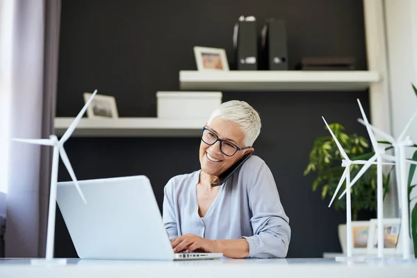 Великолепная белая старшая женщина разговаривает по телефону и печатает на ноутбуке, сидя в офисе. Концепция стартапа . — стоковое фото