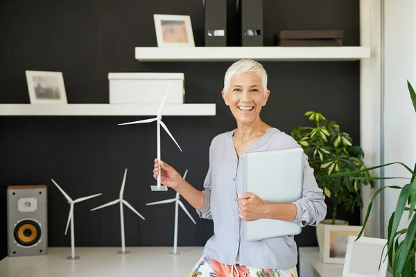 Atractiva sonriente mujer mayor caucásica vestida casual y con el pelo gris corto de pie en la oficina, sosteniendo el ordenador portátil y molino de viento. Concepto sostenible . — Foto de Stock