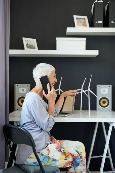 Preciosa mujer mayor caucásica hablando por teléfono y tocando el modelo de molino de viento mientras está sentado en la oficina. Concepto de creación de empresas . — Foto de Stock