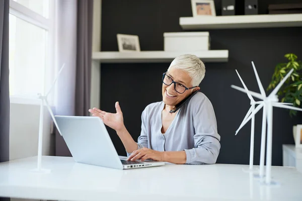 Preciosa mujer mayor caucásica hablando por teléfono y escribiendo en el ordenador portátil mientras está sentado en la oficina. Concepto de creación de empresas . — Foto de Stock