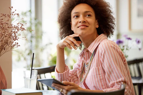 Attraktive Mischlingsfrau in gestreiftem rosa Kleid sitzt im Café und benutzt Tablet. — Stockfoto