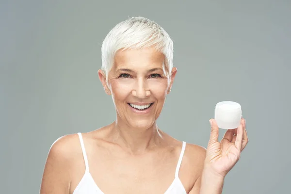 Великолепная улыбающаяся белая пожилая женщина пробует новый антивозрастной крем и смотрит в камеру. Фотография красоты . — стоковое фото