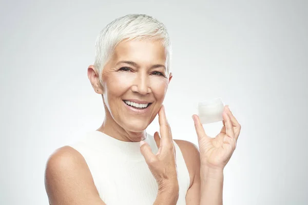 Prachtige glimlachend blanke Senior vrouw uitproberen van nieuwe anti-age crème en kijken naar de camera. Beauty fotografie. — Stockfoto